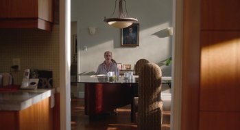 Movie still from “Beau Is Afraid” (2023), directed by Ari Aster – A man sitting at a dining room table in front of a painting; Wide shot, Over the shoulder angle