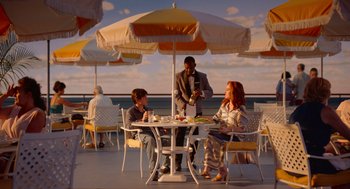 Movie still from “Beau Is Afraid” (2023), directed by Ari Aster – A man and a woman sitting at a table under an umbrella; Wide shot, High angle