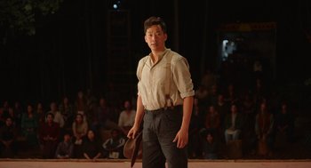 Movie still from “Beau Is Afraid” (2023), directed by Ari Aster – A young man in a white shirt holding a hat in front of an audience; Medium shot, Low angle