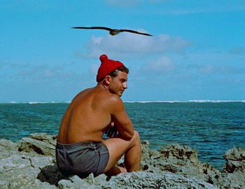Movie still from “Becoming Cousteau” (2021), directed by Liz Garbus – A man sitting on a rock near the ocean; Medium shot, Low angle