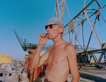 Movie still from “Becoming Cousteau” (2021), directed by Liz Garbus – A man with no shirt and no shirt eating a sandwich; Medium shot, Low angle