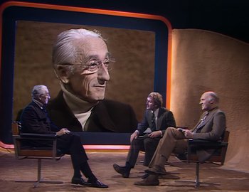 Movie still from “Becoming Cousteau” (2021), directed by Liz Garbus – A group of people sitting in front of a picture of a man; Medium shot, Over the shoulder angle