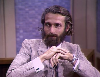 Movie still from “Becoming Cousteau” (2021), directed by Liz Garbus – A man with long hair and a goatee sitting in a chair; Close Up shot, High angle