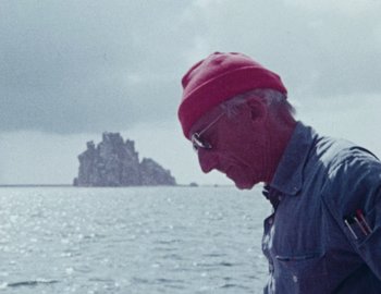Movie still from “Becoming Cousteau” (2021), directed by Liz Garbus – An older man wearing glasses and a red hat looks out to sea; Close Up shot, Low angle