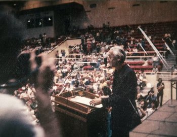 Movie still from “Becoming Cousteau” (2021), directed by Liz Garbus – An older man speaking at a podium in front of an audience; Medium shot, High angle