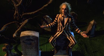 Movie still from “Beetlejuice” (1988), directed by Tim Burton – A man dressed as beetlejuice sitting on top of a tombstone; Medium shot, High angle