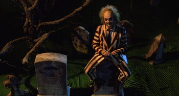Movie still from “Beetlejuice” (1988), directed by Tim Burton – A man dressed as beetlejuice sitting on top of a tombstone; Wide shot, Over the shoulder angle