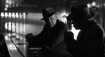 Movie still from “The Man Who Wasn't There” (2001), directed by Joel Coen – A man wearing a hat and a suit sitting at a table with another man; Medium shot, Over the shoulder angle