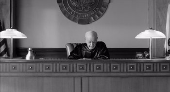 Movie still from “The Man Who Wasn't There” (2001), directed by Joel Coen – An old man sitting in a judge's chair in front of a california seal; Medium shot, Low angle