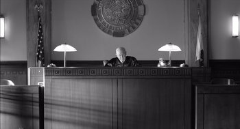 Movie still from “The Man Who Wasn't There” (2001), directed by Joel Coen – An old man sitting at a judge's desk in front of two lamps; Wide shot, High angle