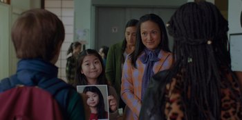 Movie still from “Better Nate Than Ever” (2022), directed by Tim Federle – A woman holding a picture of a young girl in front of a group of people; Medium shot, Over the shoulder angle