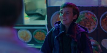 Movie still from “Better Nate Than Ever” (2022), directed by Tim Federle – A young boy smiles brightly in a dark room; Close Up shot, Over the shoulder angle