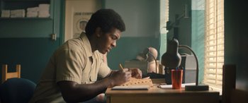 Movie still from “Big George Foreman” (2023), directed by George Tillman Jr. – A man sitting at a desk writing on a piece of paper; Medium shot, Low angle