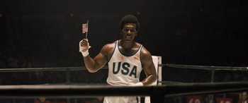 Movie still from “Big George Foreman” (2023), directed by George Tillman Jr. – A man holding an american flag in a boxing ring; Medium shot, Low angle
