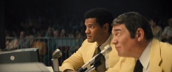 Movie still from “Big George Foreman” (2023), directed by George Tillman Jr. – A man in a yellow suit sitting next to a microphone; Medium shot, Low angle