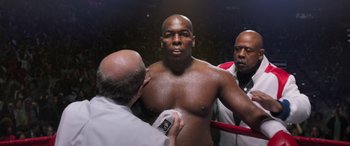 Movie still from “Big George Foreman” (2023), directed by George Tillman Jr. – A man in a boxing ring getting his chest examined by a doctor; Medium shot, Low angle
