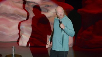 Movie still from “Bill Burr: Live at Red Rocks” (2022), directed by Mike Binder – A bald man in a blue shirt is holding a microphone; Medium shot, High angle