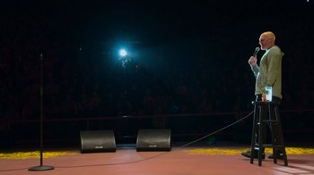 Movie still from “Bill Burr: Live at Red Rocks” (2022), directed by Mike Binder – A stage with two speakers and a microphone in front of a crowd of people; Extreme Wide shot, High angle