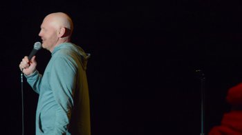 Movie still from “Bill Burr: Live at Red Rocks” (2022), directed by Mike Binder – A bald man in a blue shirt is standing in the dark; Medium shot, Low angle