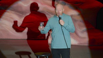 Movie still from “Bill Burr: Live at Red Rocks” (2022), directed by Mike Binder – A man holding a glass of water while standing on a stage; Medium shot, Over the shoulder angle