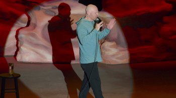 Movie still from “Bill Burr: Live at Red Rocks” (2022), directed by Mike Binder – A bald man in a blue shirt is holding a microphone; Wide shot, High angle