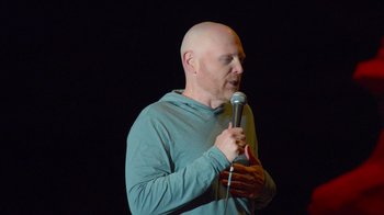 Movie still from “Bill Burr: Live at Red Rocks” (2022), directed by Mike Binder – A bald man holding a microphone in front of a black background; Medium shot, Low angle