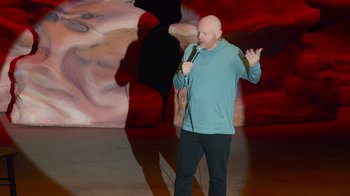Movie still from “Bill Burr: Live at Red Rocks” (2022), directed by Mike Binder – A bald man in a blue shirt is holding a microphone; Medium shot, High angle