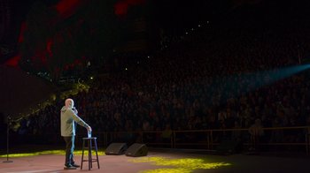Movie still from “Bill Burr: Live at Red Rocks” (2022), directed by Mike Binder – A man standing on a stool in front of an audience; Extreme Wide shot, High angle