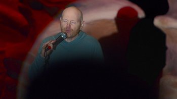 Movie still from “Bill Burr: Live at Red Rocks” (2022), directed by Mike Binder – A man with a bald head is singing into a microphone; Medium shot, Overhead angle