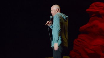 Movie still from “Bill Burr: Live at Red Rocks” (2022), directed by Mike Binder – A bald man standing on a stage holding a microphone; Medium shot, Over the shoulder angle
