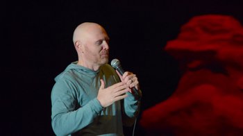 Movie still from “Bill Burr: Live at Red Rocks” (2022), directed by Mike Binder – A bald man holding a microphone while standing on a stage; Medium shot, Low angle
