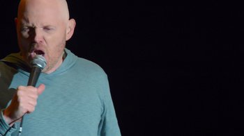Movie still from “Bill Burr: Live at Red Rocks” (2022), directed by Mike Binder – A bald man wearing a blue shirt is standing in front of a black background; Close Up shot, Low angle