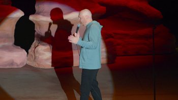 Movie still from “Bill Burr: Live at Red Rocks” (2022), directed by Mike Binder – A bald man in a blue jacket is holding a microphone; Wide shot, High angle