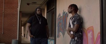 Movie still from “Black as Night” (2021), directed by Maritte Lee Go – Two men standing next to each other in front of a building; Medium shot, Low angle