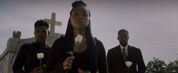 Movie still from “Black as Night” (2021), directed by Maritte Lee Go – A woman holding a white flower in front of a cross; Medium shot, Low angle