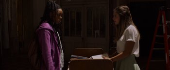 Movie still from “Black as Night” (2021), directed by Maritte Lee Go – Two young women are standing in front of a stack of books; Medium shot, Over the shoulder angle