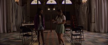 Movie still from “Black as Night” (2021), directed by Maritte Lee Go – Two young women are standing in a room with chairs; Medium shot, Low angle