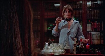 Movie still from “Black Christmas” (1974), directed by Bob Clark – A woman standing in front of a table holding a drink; Medium shot, Over the shoulder angle