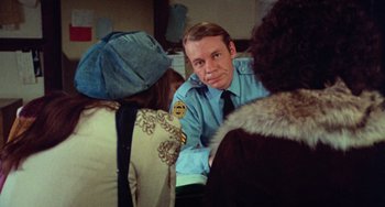 Movie still from “Black Christmas” (1974), directed by Bob Clark – A man in a police uniform sitting in front of a group of people; Close Up shot, Over the shoulder angle