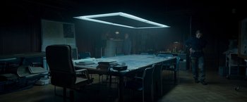 Movie still from “Black Crab” (2022), directed by Adam Berg – A large table in a dark room with chairs around it; Wide shot, High angle