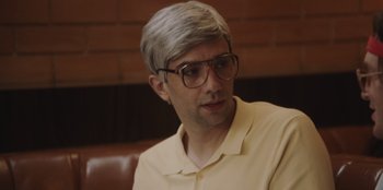 Movie still from “BlackBerry” (2023), directed by Matt Johnson – A man with grey hair wearing glasses sitting in front of a brick wall; Close Up shot, Over the shoulder angle