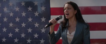 Movie still from “Blacklight” (2022), directed by Mark Williams – A woman holding a microphone in front of an american flag; Medium shot, Low angle