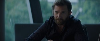 Movie still from “Blacklight” (2022), directed by Mark Williams – A man with a goatee sitting in front of a window; Close Up shot, Over the shoulder angle
