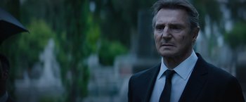 Movie still from “Blacklight” (2022), directed by Mark Williams – A man wearing a suit and tie standing in front of trees; Close Up shot, Over the shoulder angle