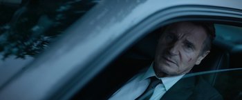Movie still from “Blacklight” (2022), directed by Mark Williams – A person wearing a suit driving a car; Close Up shot, High angle
