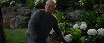 Movie still from “Blacklight” (2022), directed by Mark Williams – An older man wearing a sweater and gloves in a garden; Close Up shot, Low angle