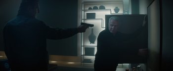 Movie still from “Blacklight” (2022), directed by Mark Williams – A man holding a gun in front of another man in a dark room; Medium shot, Over the shoulder angle