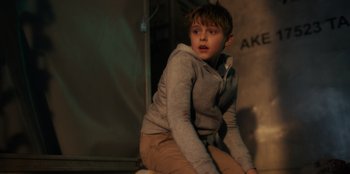 Movie still from “Blood Red Sky” (2021), directed by Peter Thorwarth – A young boy sitting on the ground in front of a wall; Medium shot, Low angle