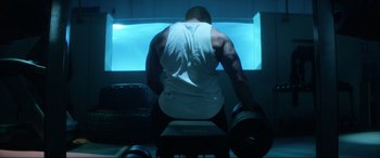 Movie still from “Bloodshot” (2020), directed by Dave Wilson – A man in a white tank top is lifting a dumbbell; Medium shot, Over the shoulder angle