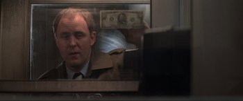 Movie still from “Blow Out” (1981), directed by Brian De Palma – A man in a suit looking at a dollar bill through a window; Close Up shot, Over the shoulder angle